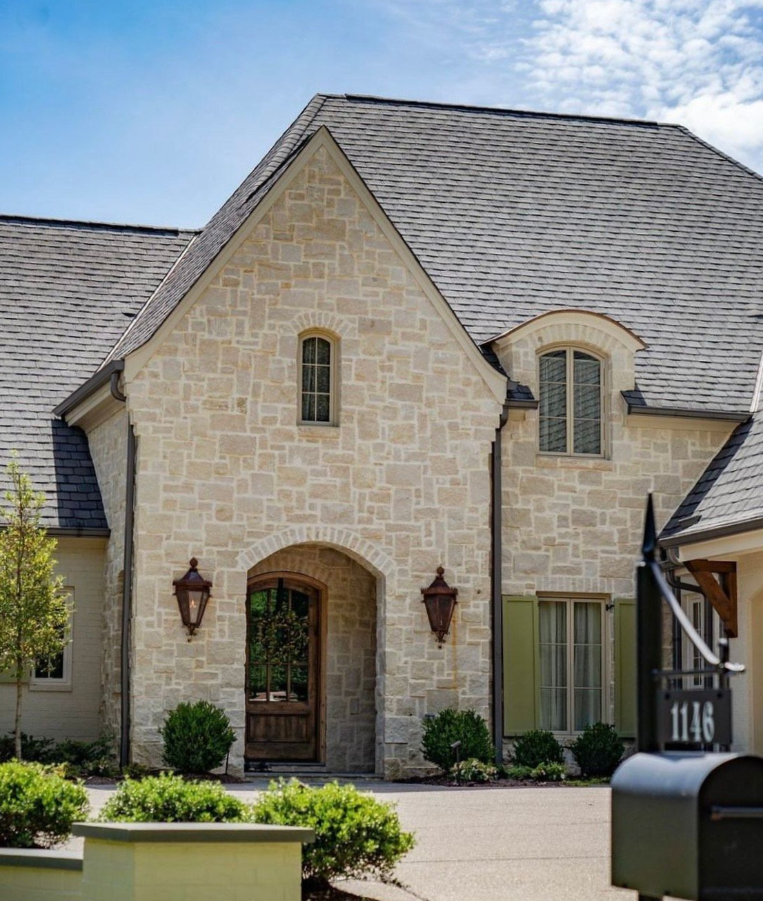Stone-clad house with arched entryway, small windows, green shutters, and a gray shingle roof.