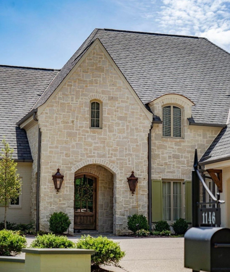 Stone-clad house with arched entryway, small windows, green shutters, and a gray shingle roof.