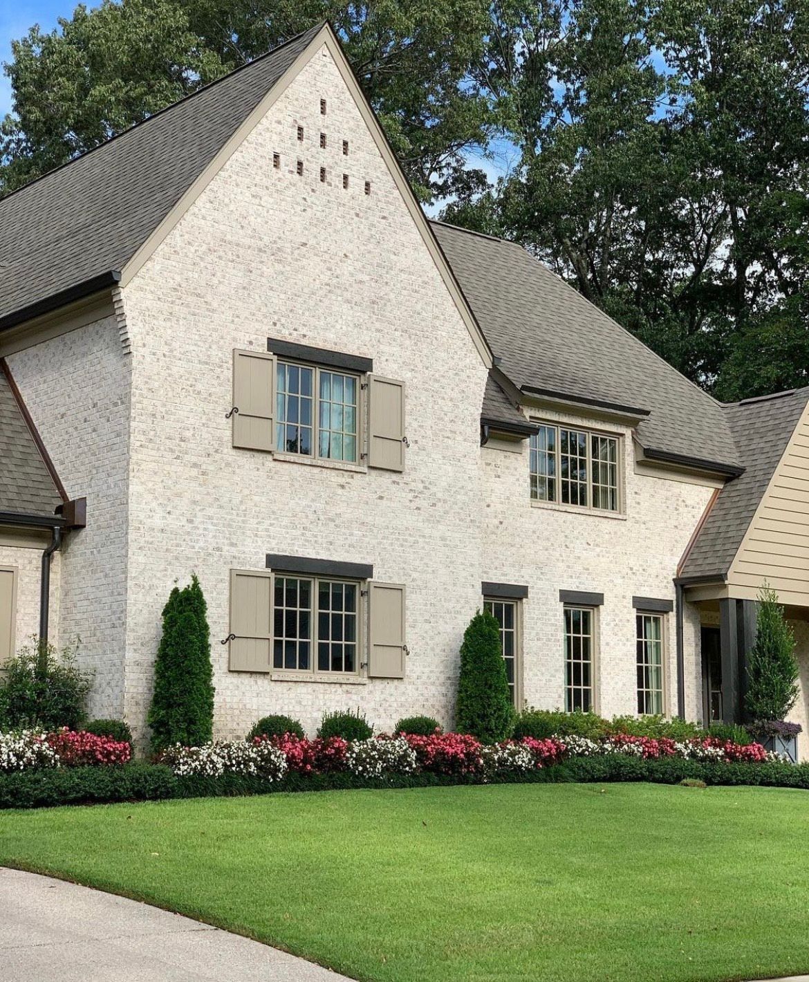 Two-story house with light stucco walls, gray roof, beige window shutters, and a well-manicured lawn and garden.