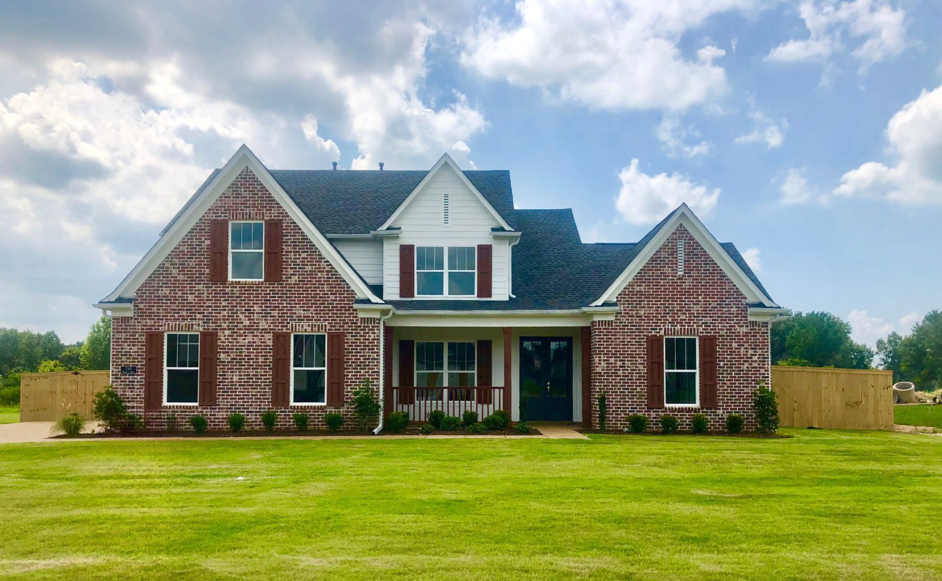 Brick house with dark roof and red shutters, set on a green lawn with a partly cloudy sky.