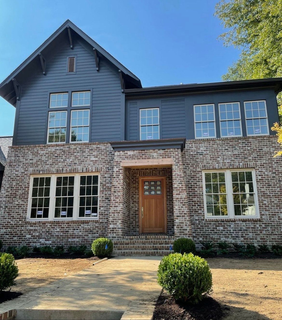 Two-story house with blue-gray siding, brick exterior, and a wooden front door, set on a sunny day.