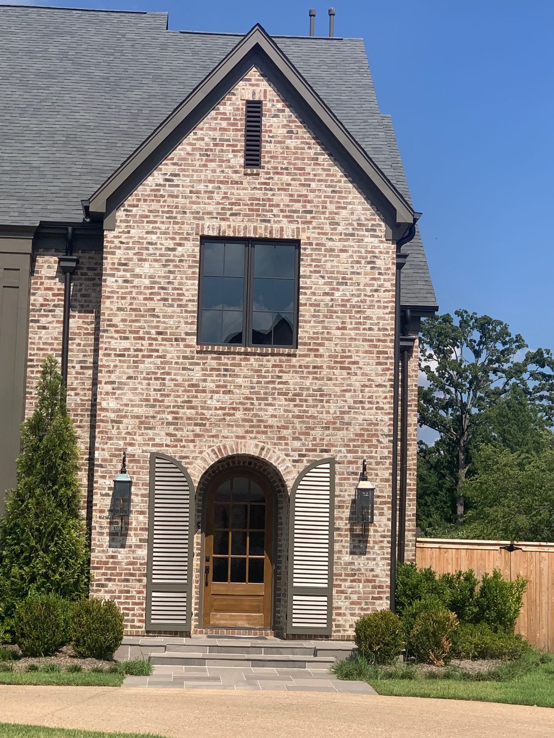 Brick house with arched doorway, shutters, and dark trim. Grey roof, blue sky.