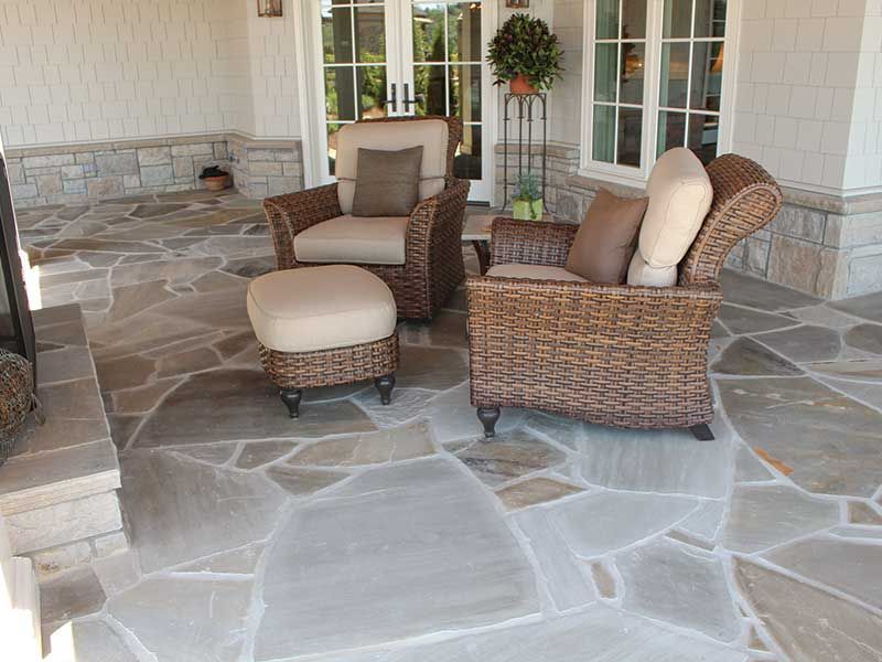 Wicker patio furniture with cushions on a stone patio, beside a fireplace and double doors.