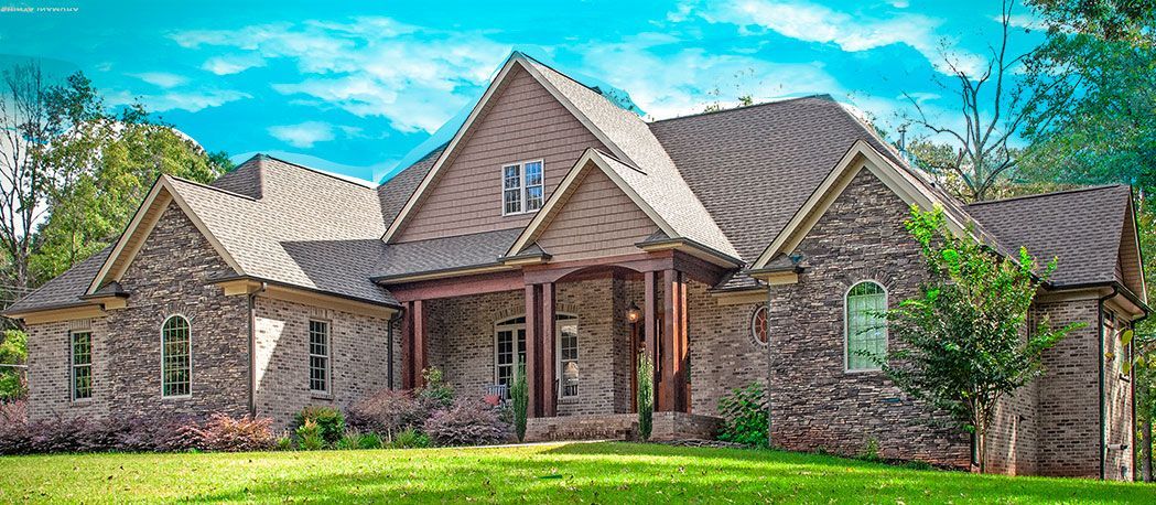 Stone-walled house with multiple gables under a bright blue sky. Green lawn and trees surround the structure.