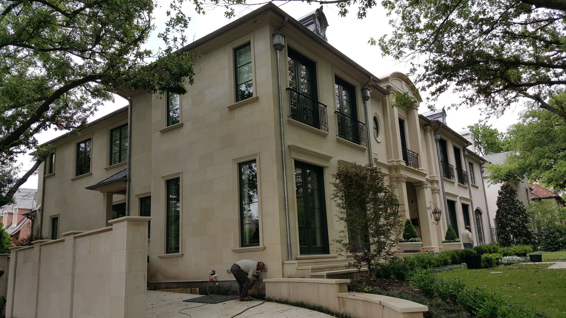 Beige two-story house with dark window frames, stairs, and a low wall; trees frame the view.
