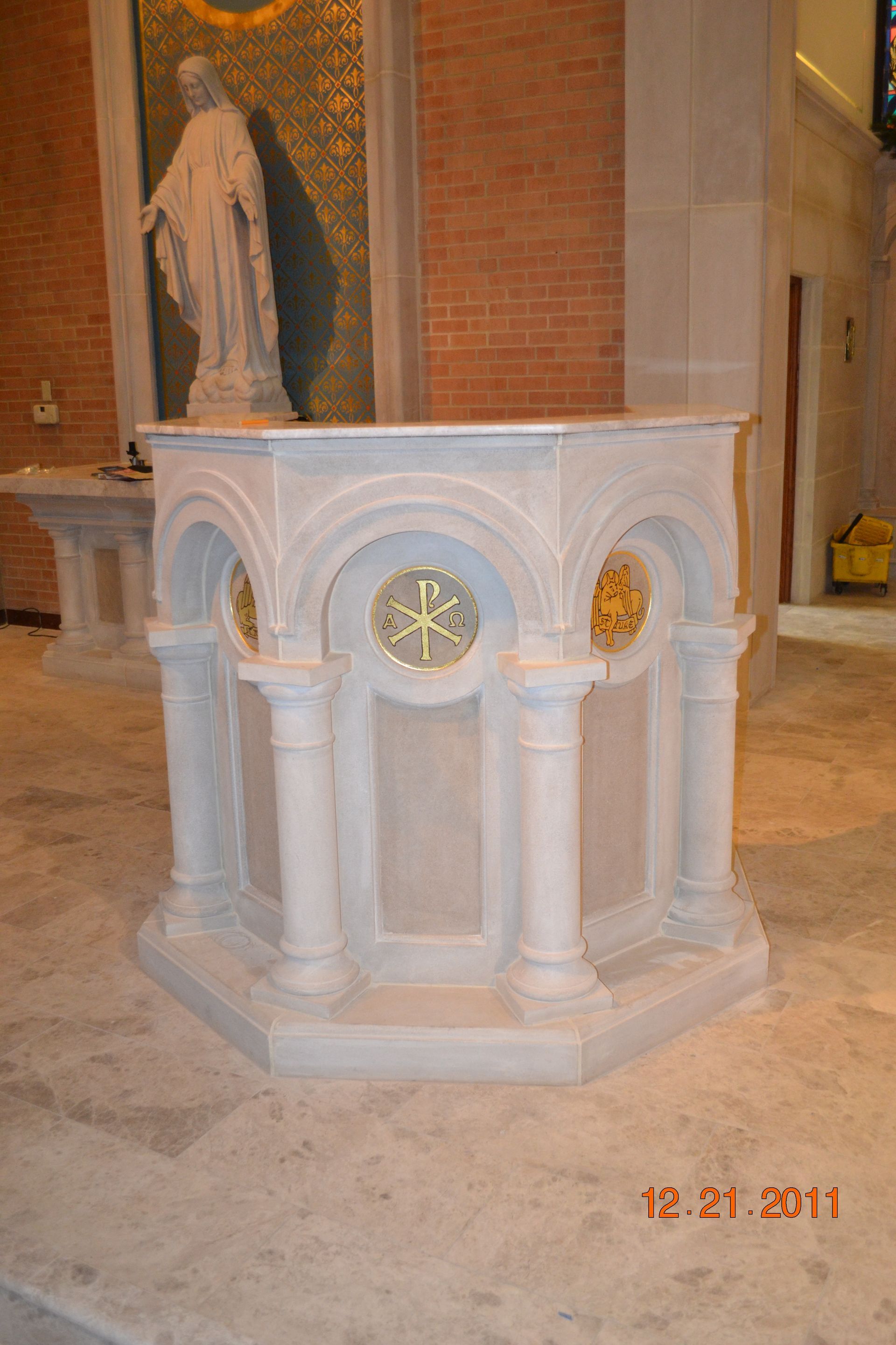 Stone baptismal font with gold accents and a Chi Rho symbol, inside a church.