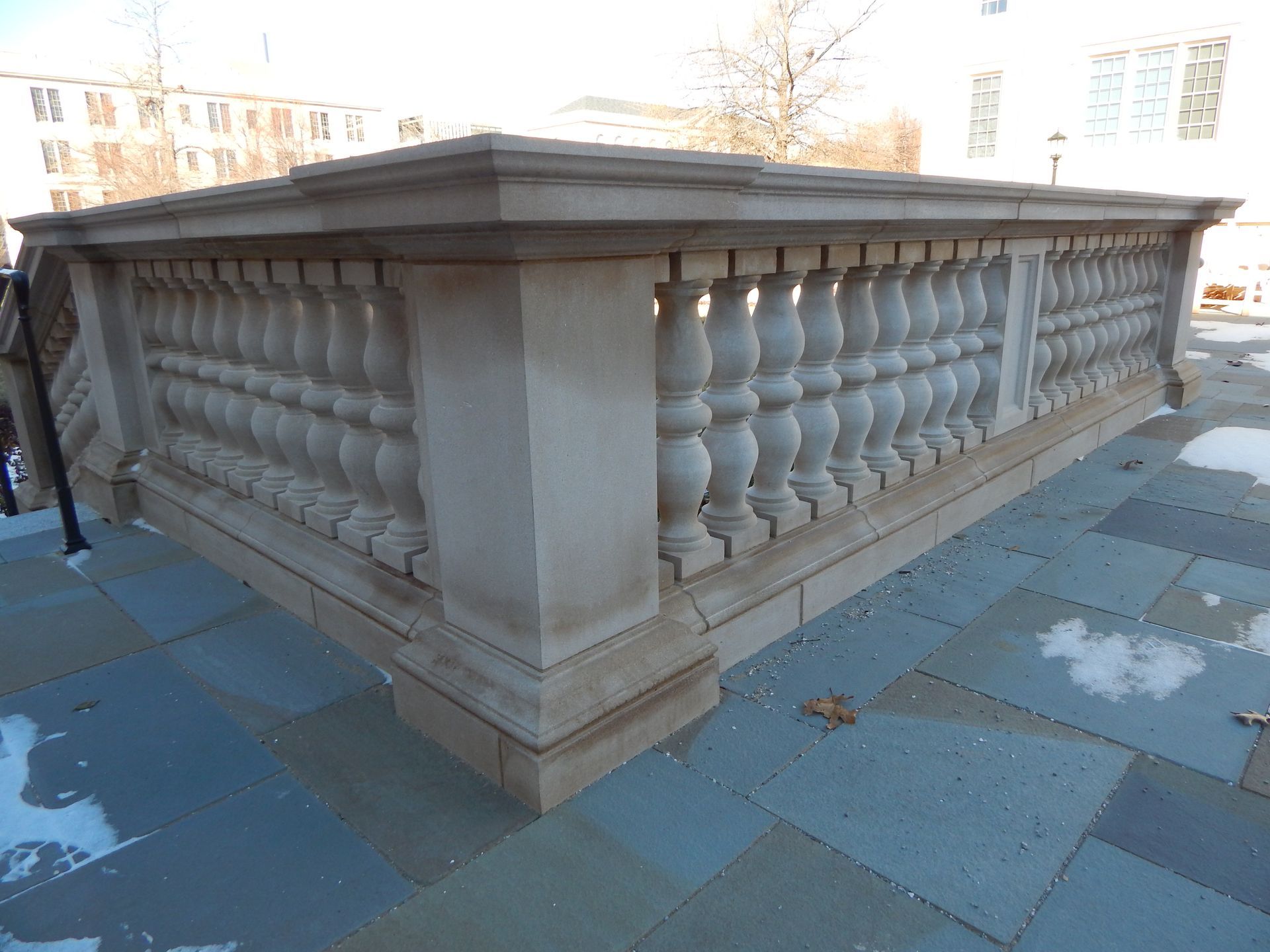 Stone balustrade on a paved surface, with ornate balusters. Sunlight, snow patches.
