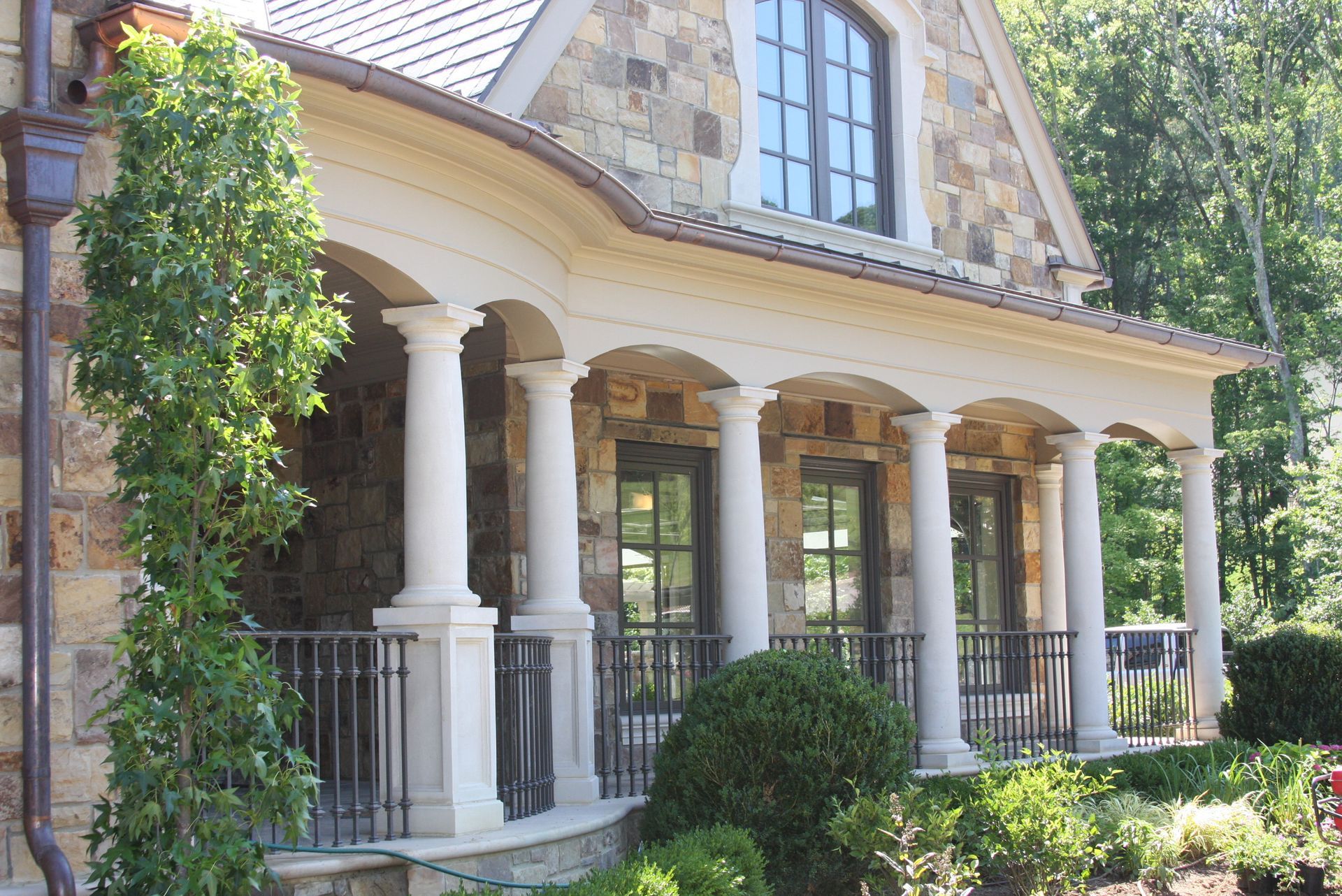 Stone house with porch featuring columns, black railing, and lush greenery.