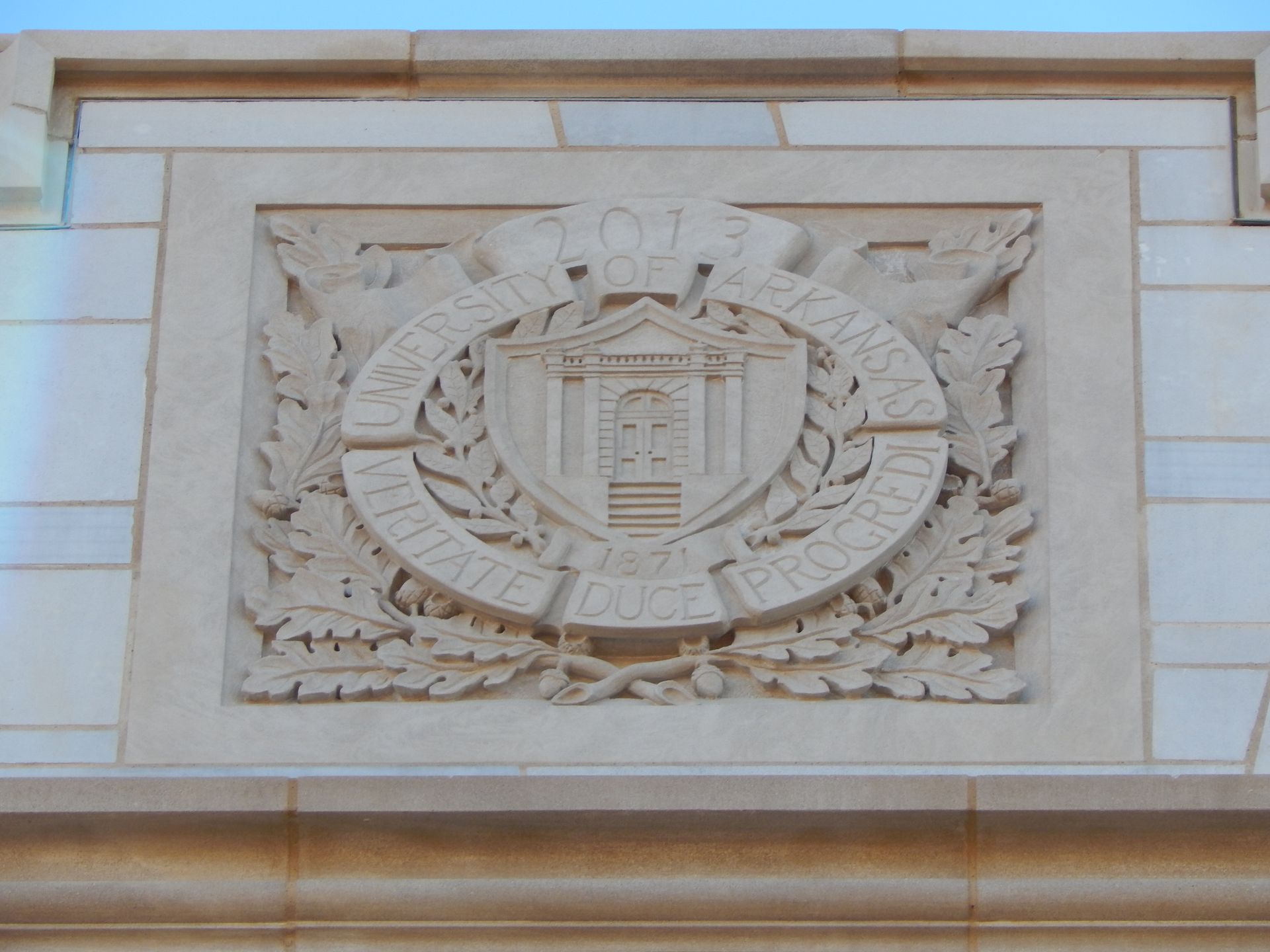 Stone carving: a building facade emblem featuring a central building image, surrounded by a wreath and inscription.