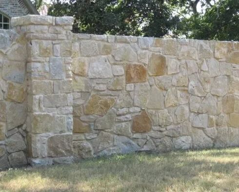 Stone wall with a pillar, built with light-colored, irregularly shaped rocks, on a grassy lawn.