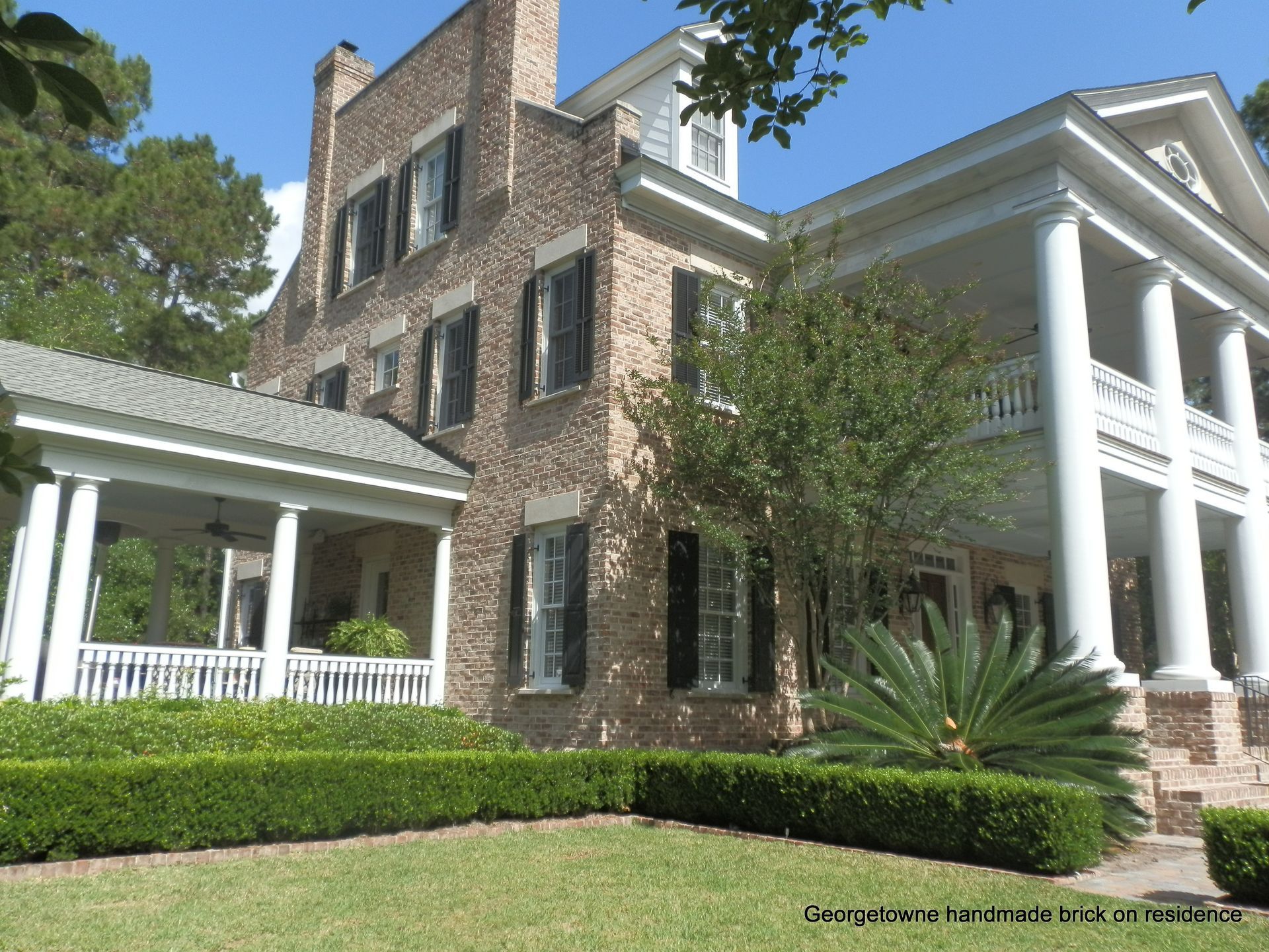 Two-story brick mansion with white columns, shutters, porch, and well-manicured lawn and hedges.