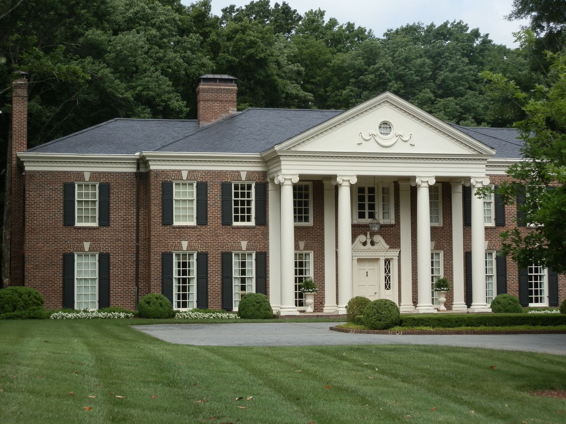 Grand brick house with white columns, shutters, and a well-manicured lawn, set against a backdrop of trees.