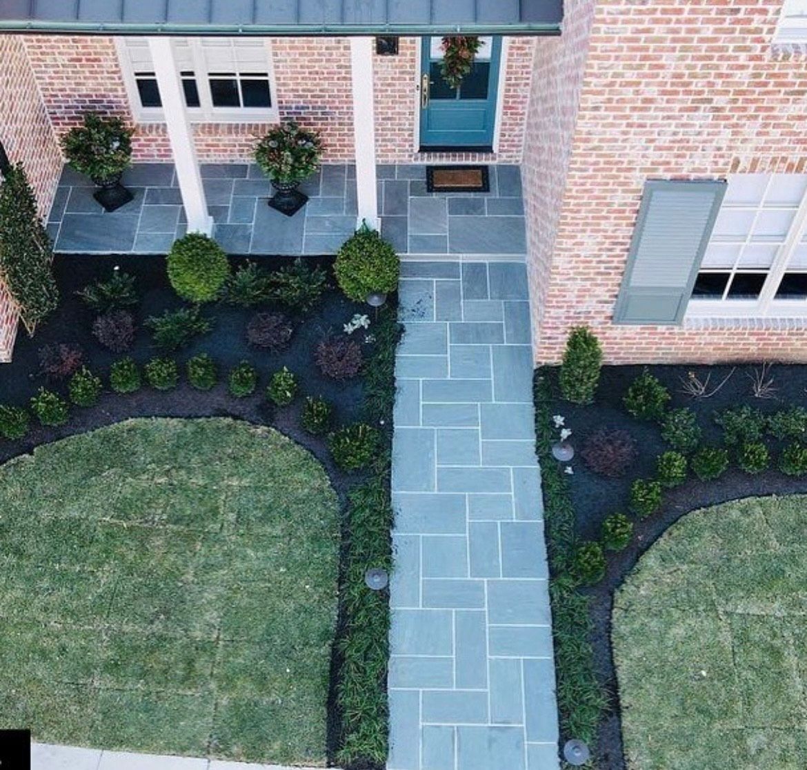 Brick house with teal door and blue-gray stone walkway, flanked by manicured landscaping and green lawn.
