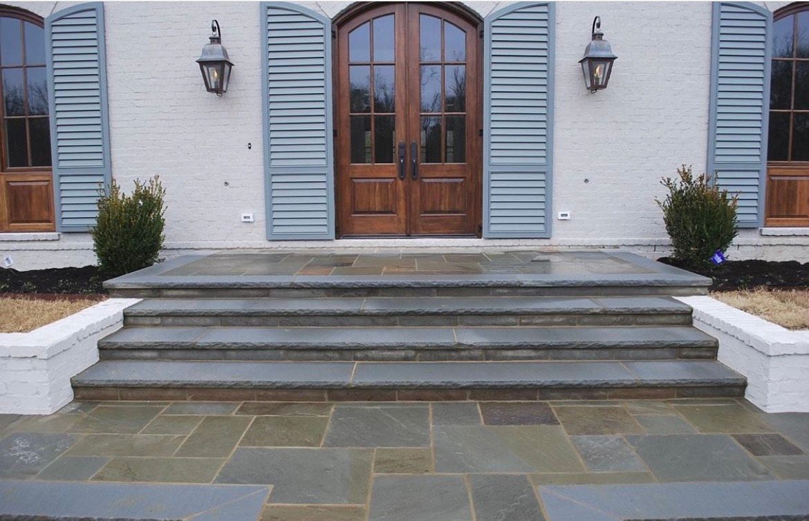 Exterior view of a house entrance with stone steps, wooden double doors, and blue shutters.