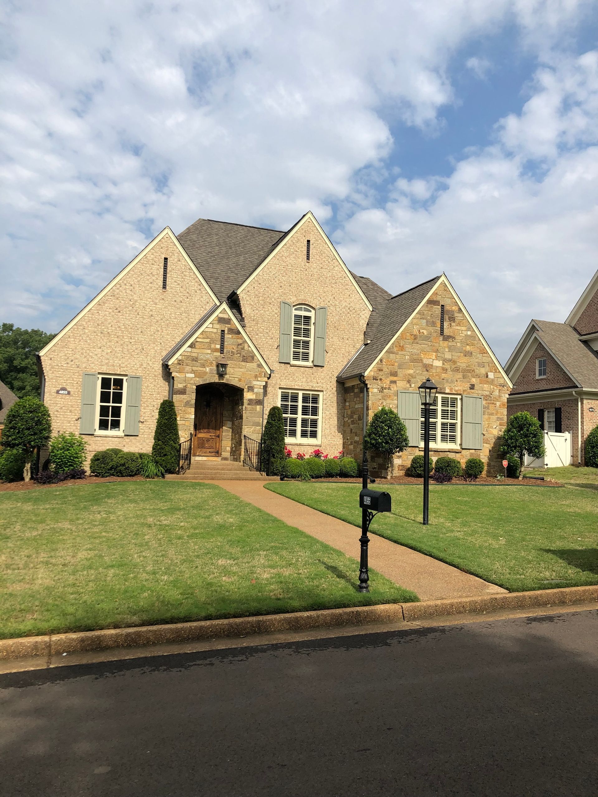 Stone house with light green shutters, brown roof, and a green lawn on a sunny day.