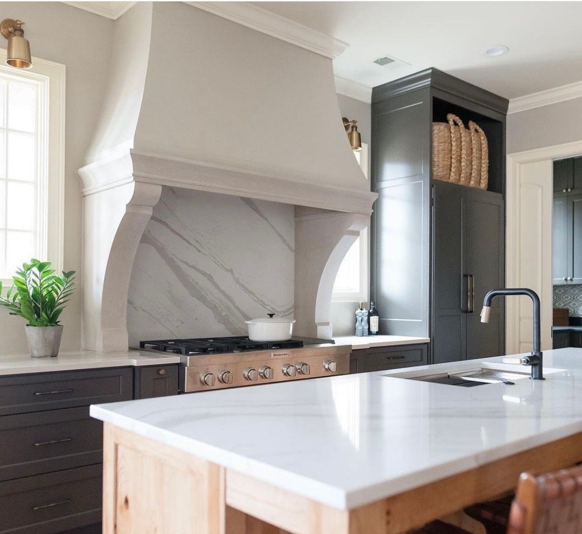 Kitchen with a white marble island, stove, and range hood, dark cabinets, and a tall, dark storage cabinet.
