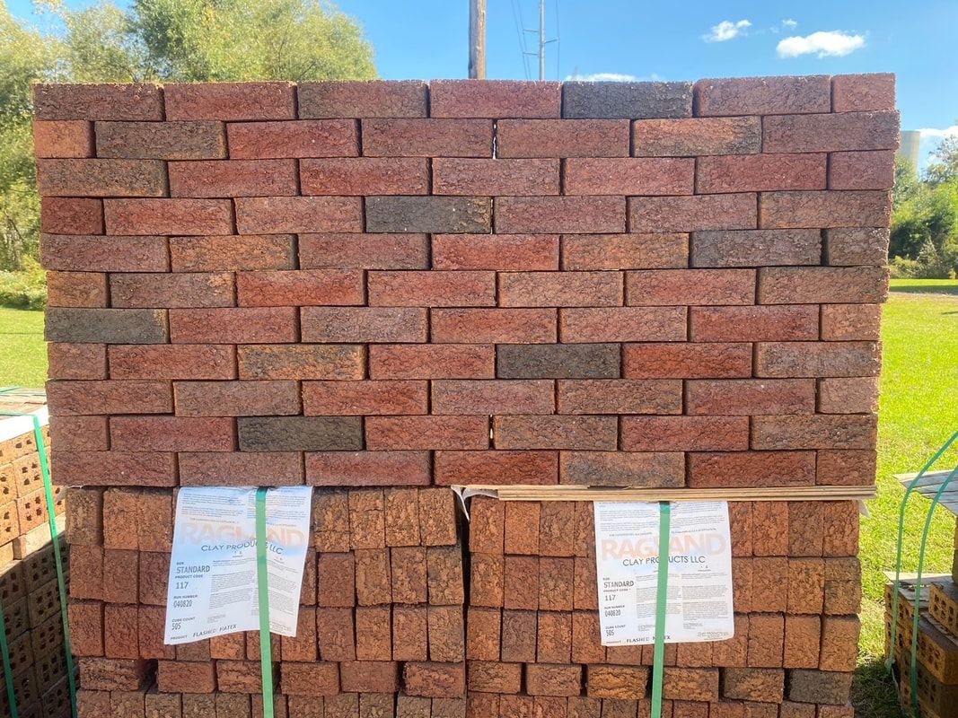 Stack of red-brown bricks, bound with green straps, in an outdoor setting with green grass and trees.