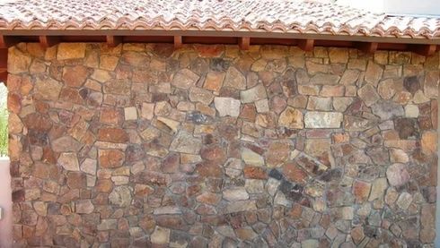 Stone wall with reddish-brown irregular shaped stones, under a terracotta tile roof with wooden beams.