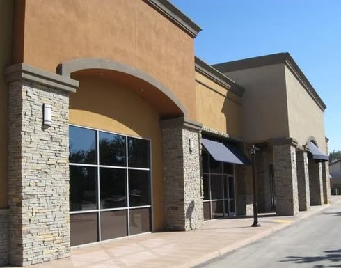 Empty retail storefront with stone pillars, tinted windows, and awnings under a blue sky.