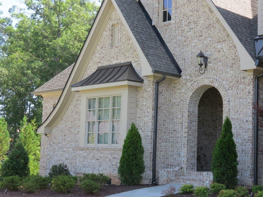 Beige brick house with arched doorway, black roof, and manicured landscaping.