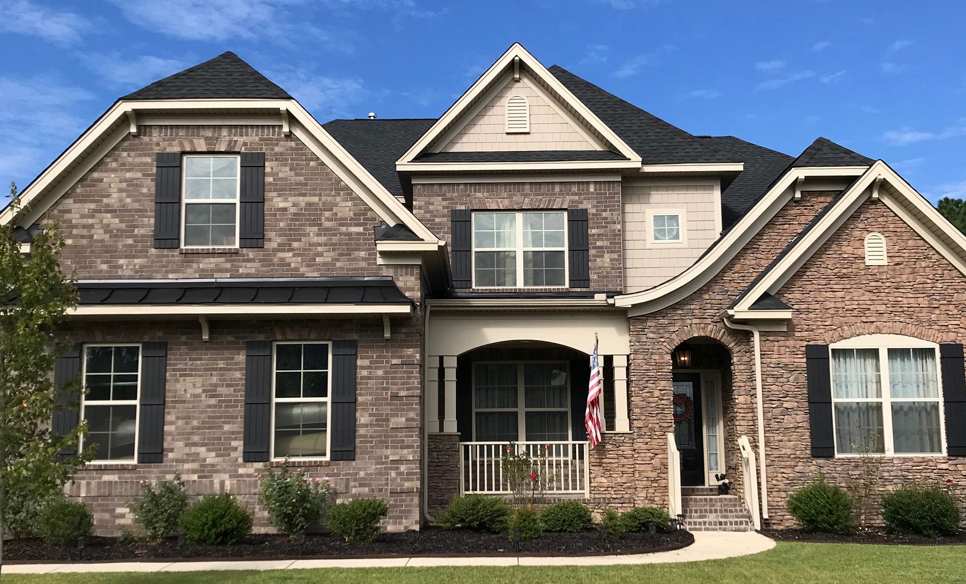 Brick-faced, two-story house with dark shutters, front porch, and flag against a blue sky.