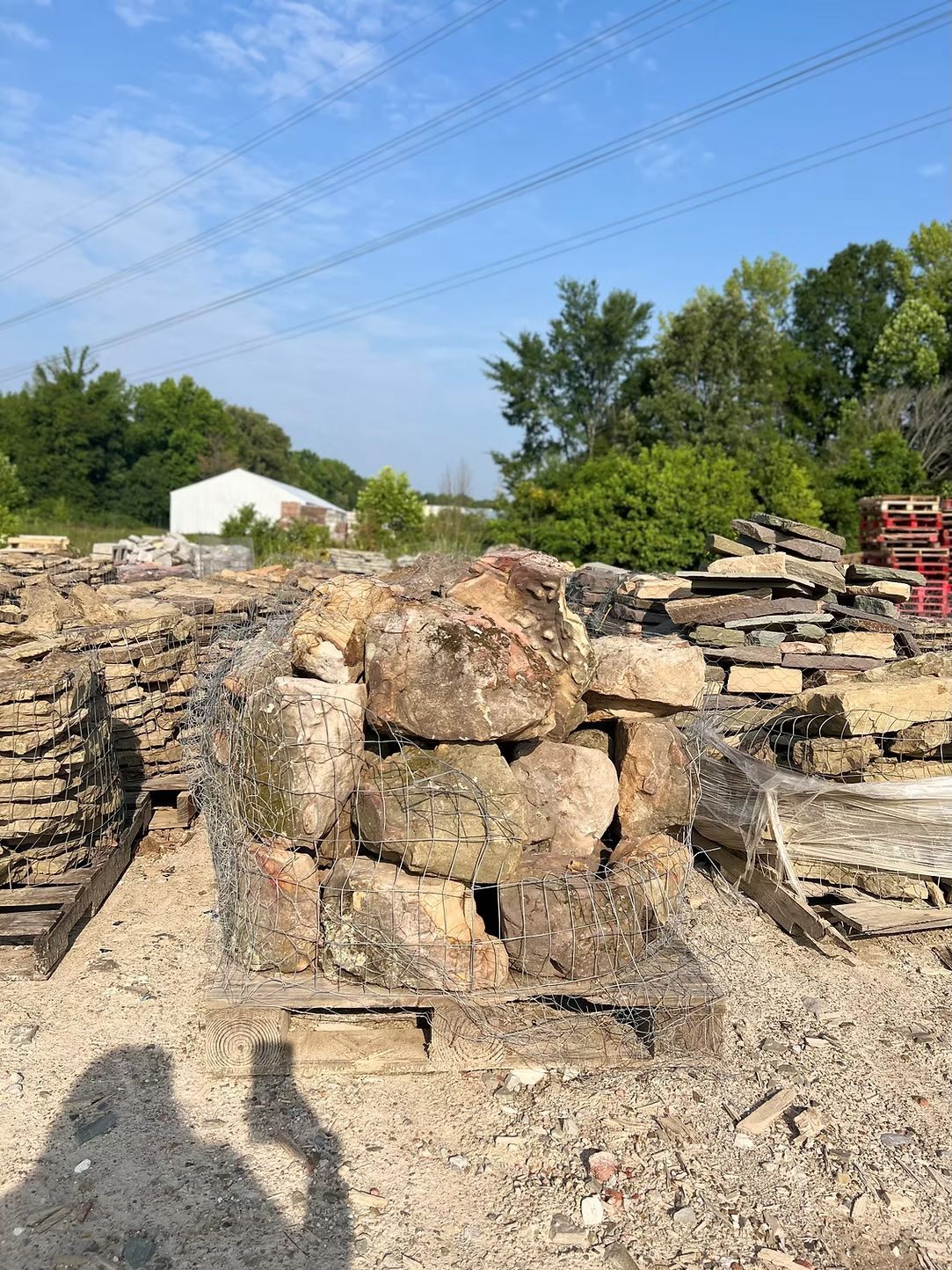 Large, weathered rocks in a gravel lot with stacks of flat stones. Trees and a building in the background.