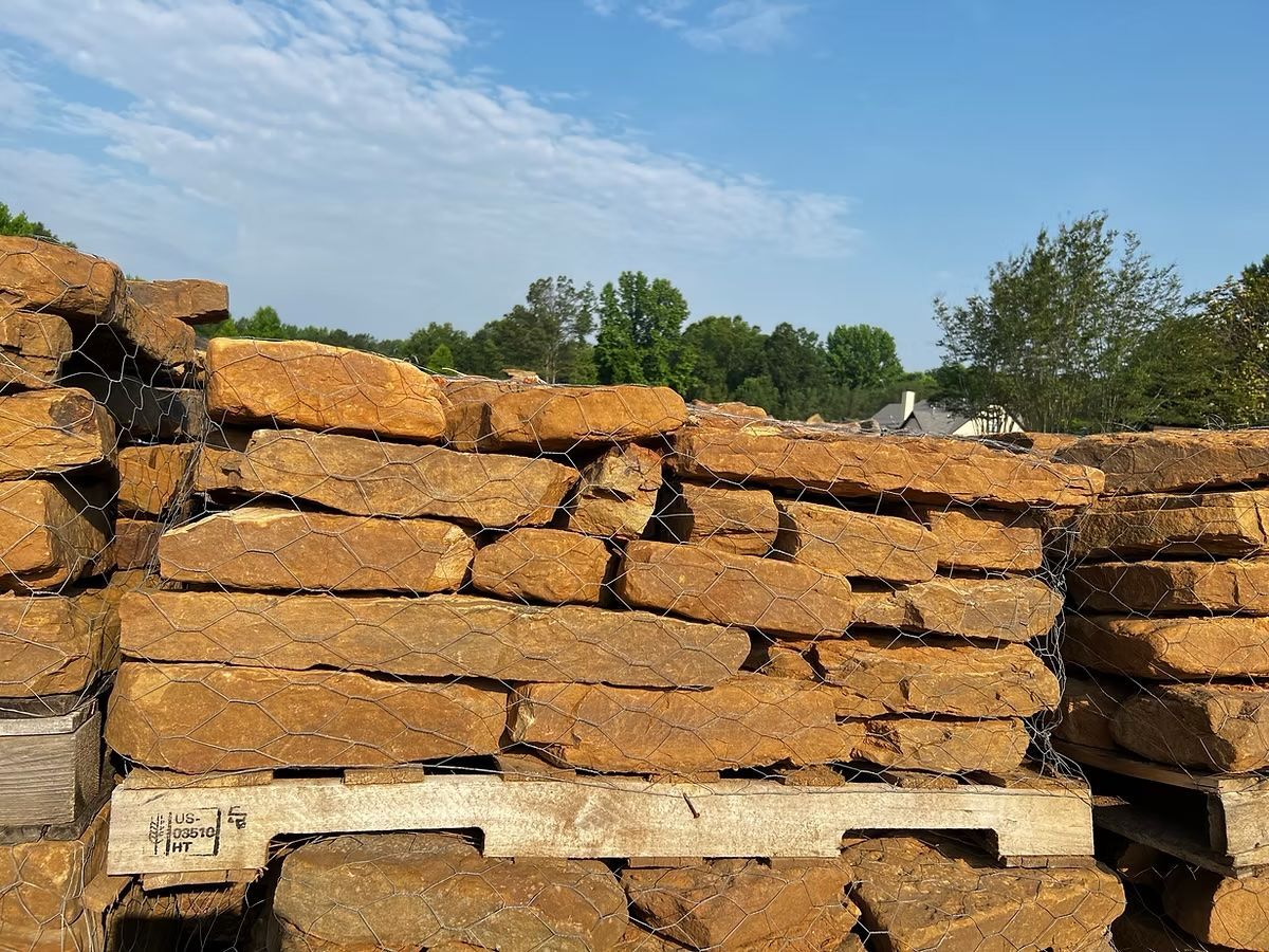 Stacked tan-colored stone pavers on a pallet, under a blue sky, trees in the background.