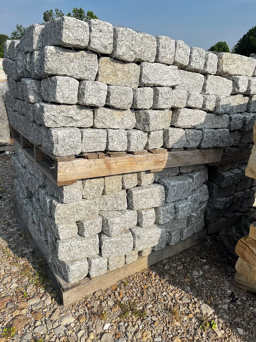 Stacked gray granite cobblestones on a wooden pallet outdoors under a blue sky.