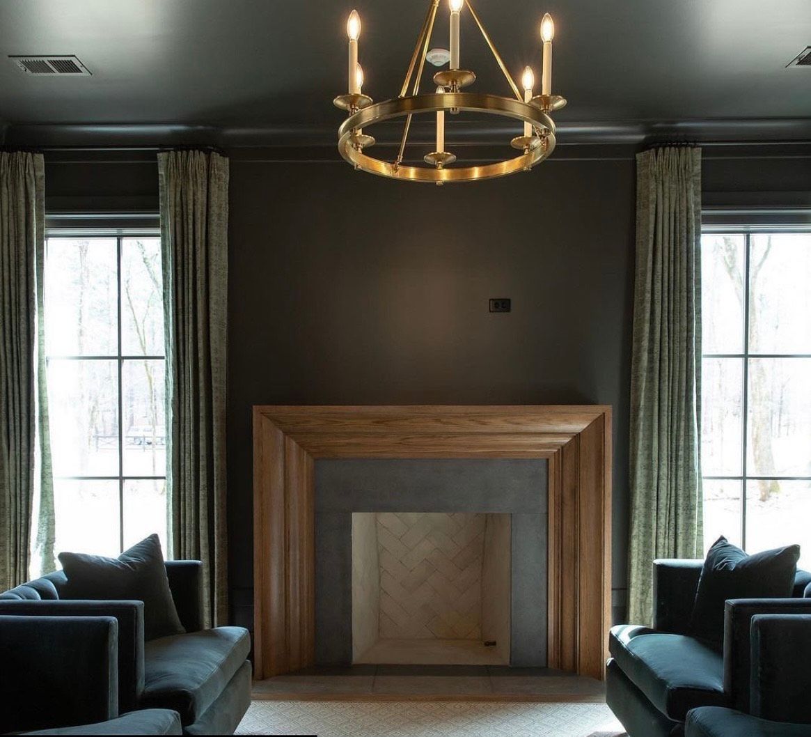 Dark-walled living room with fireplace, two armchairs, windows with curtains, and a gold chandelier.