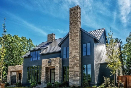Modern gray house with stone accents, tall chimney, and large windows against a blue sky.