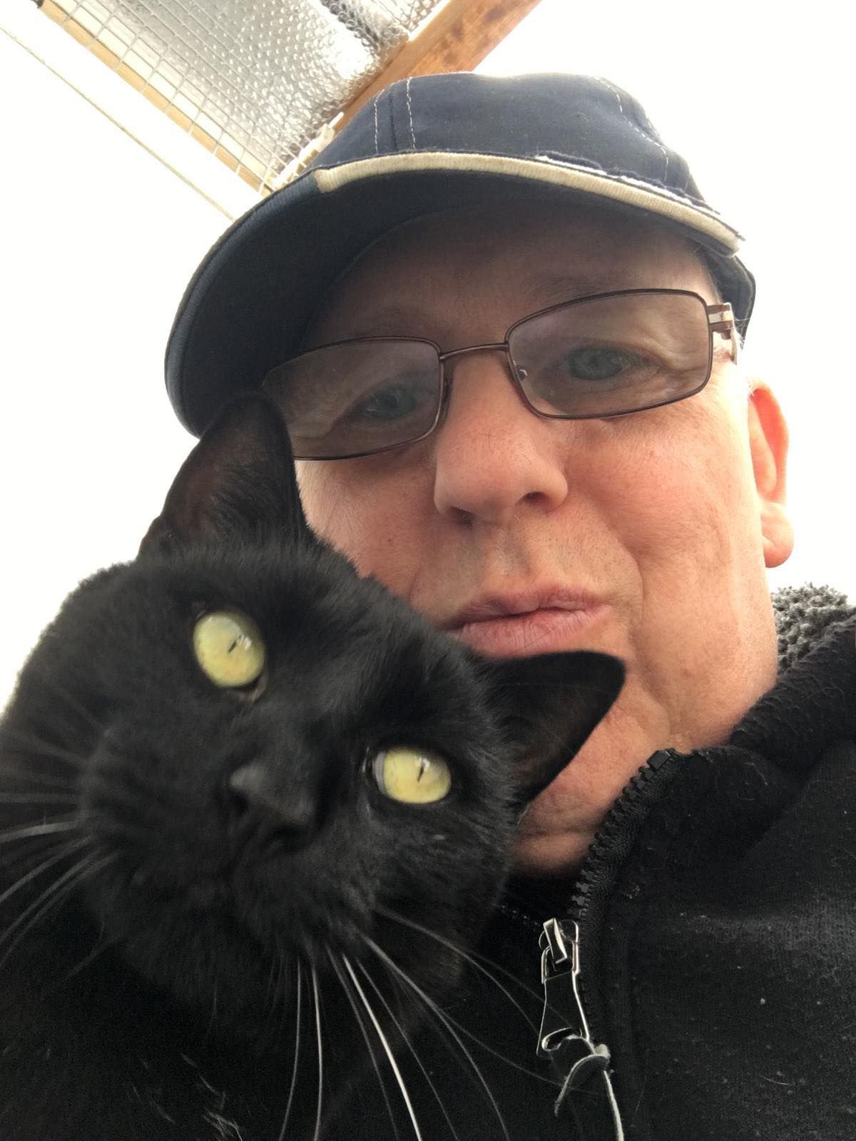 A man wearing glasses and a hat is holding a black cat