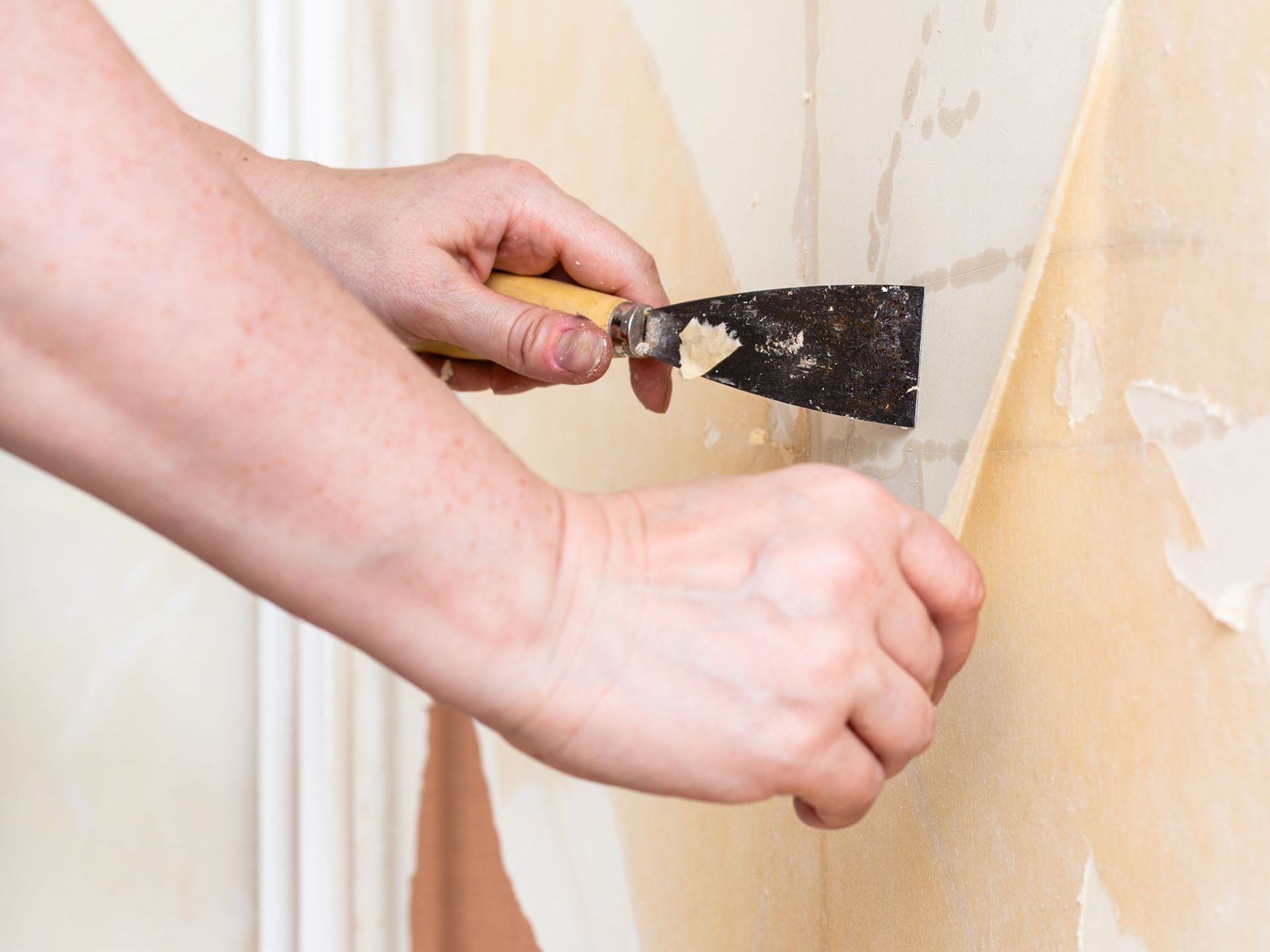 A person is removing wallpaper from a wall with a spatula.