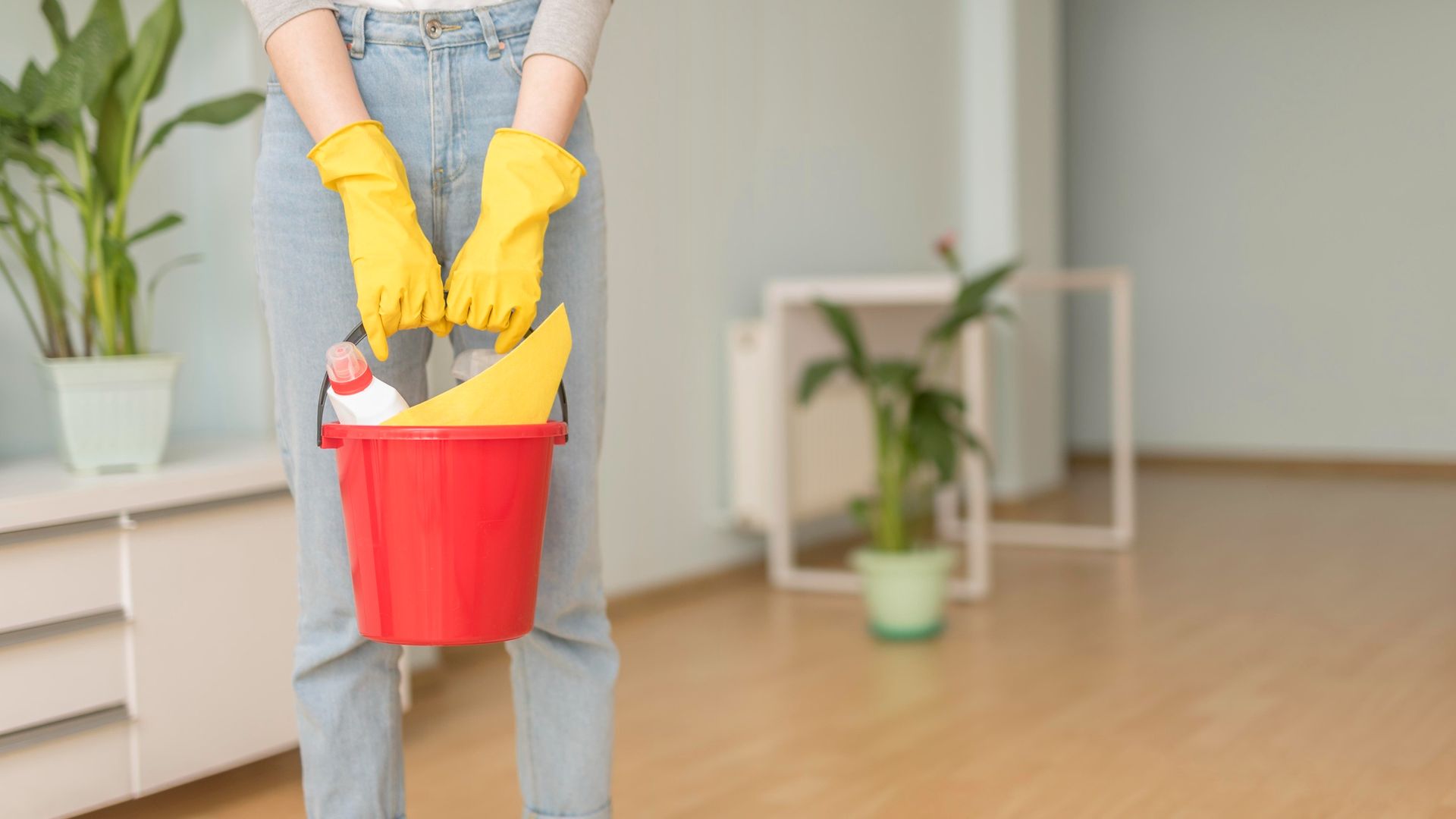 Person in yellow gloves holding a red bucket with cleaning supplies in a room.