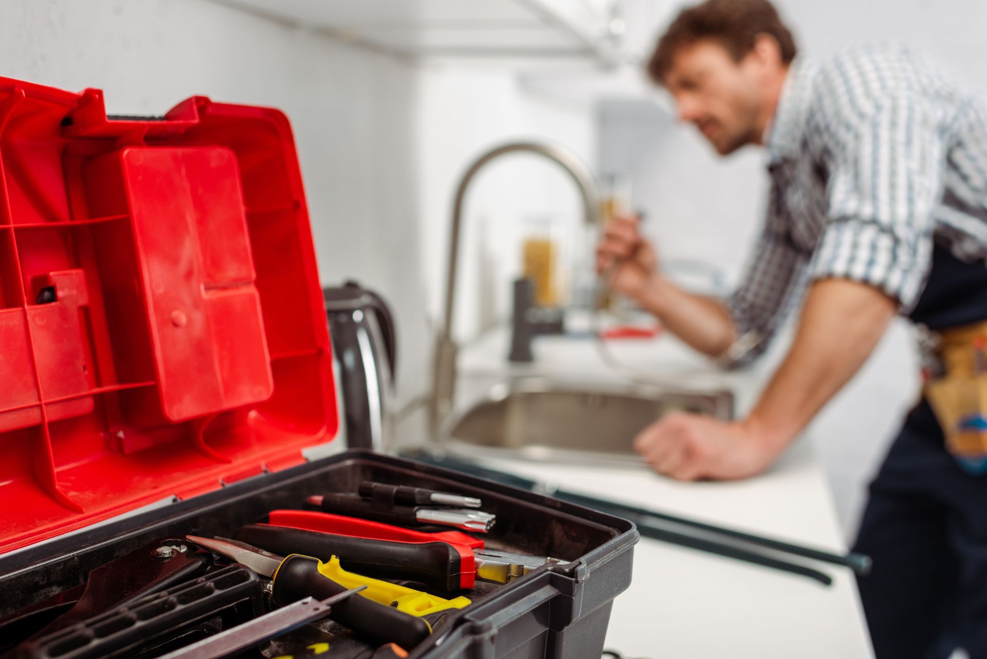 Red toolbox with tools in foreground, plumber examining kitchen faucet in background.