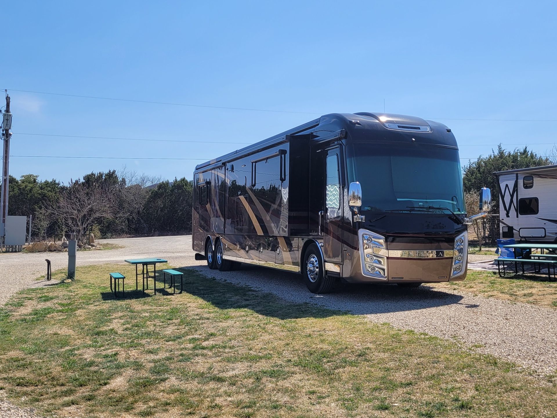 A large rv is parked in a gravel lot next to a picnic table.