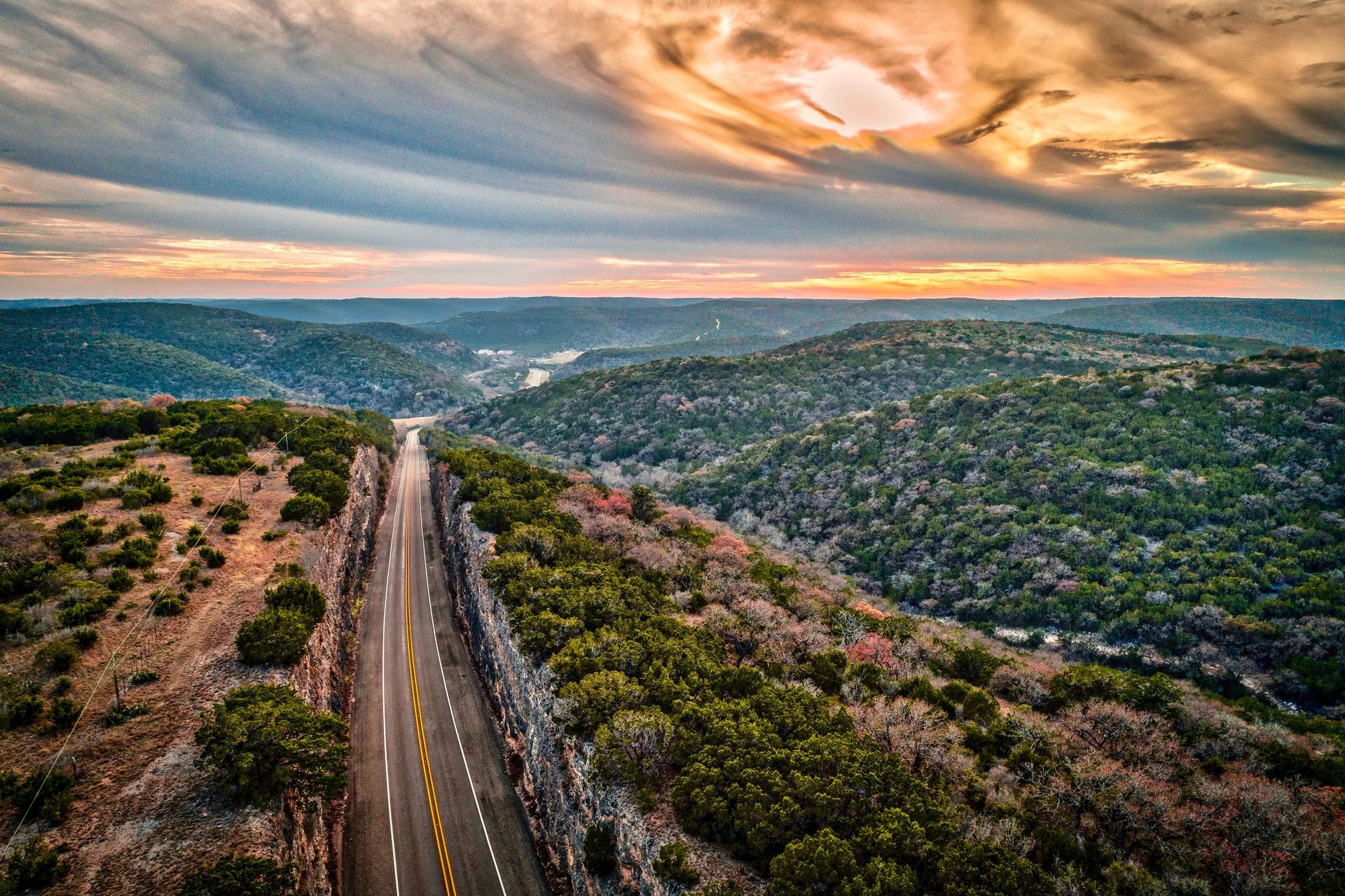 An aerial view of a road going through a valley surrounded by trees at sunset.