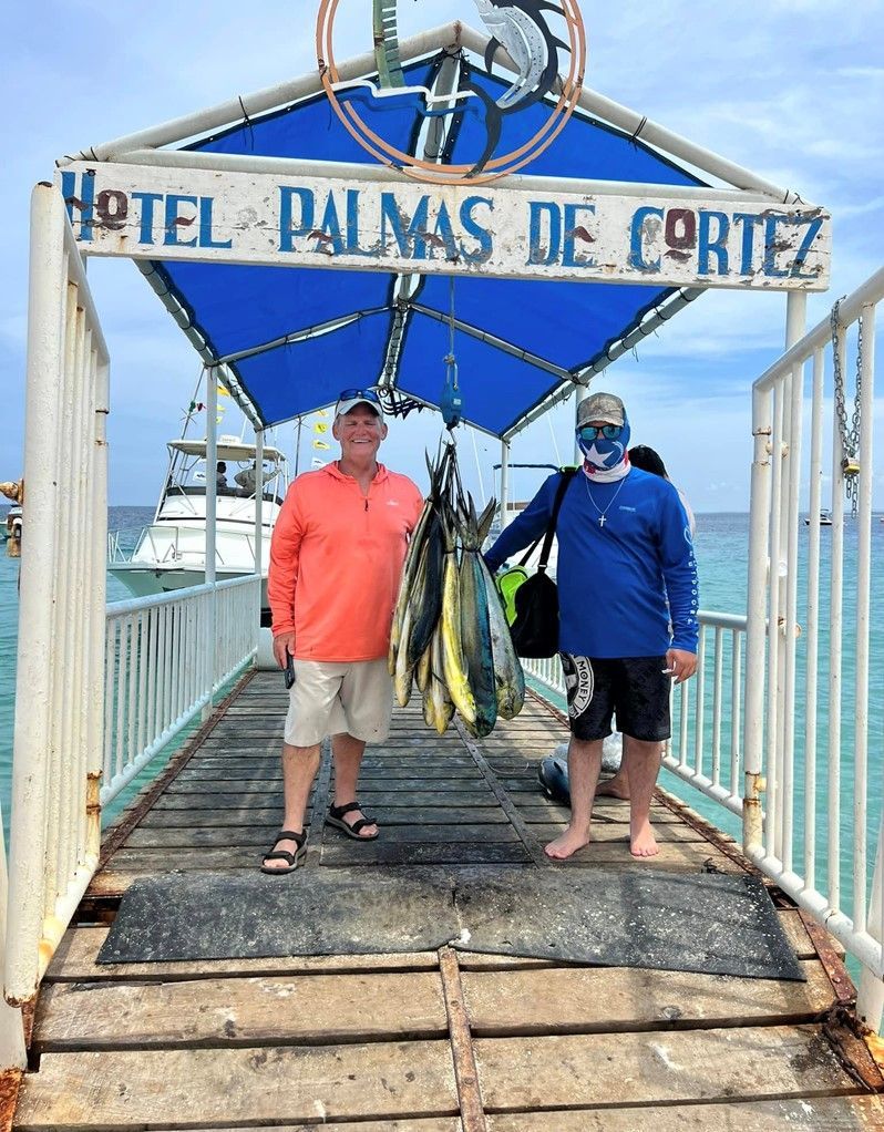 two men standing on a dock under a sign that says hotel palmas de cortez holding a tuna catch