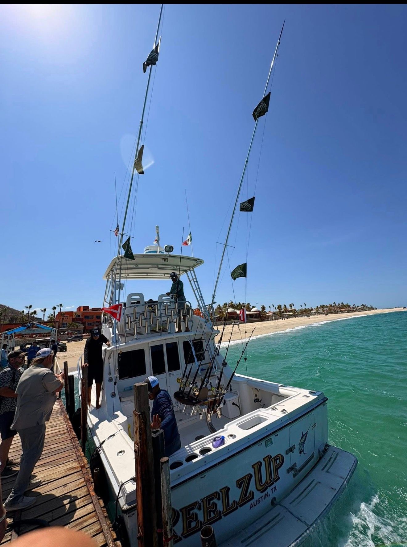 Fishing boat docked by a beach under a clear blue sky, with anglers on the pier.