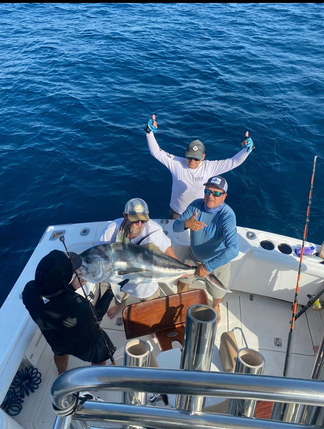 Three anglers on a boat posing with a large fish on the deck, blue ocean behind them.