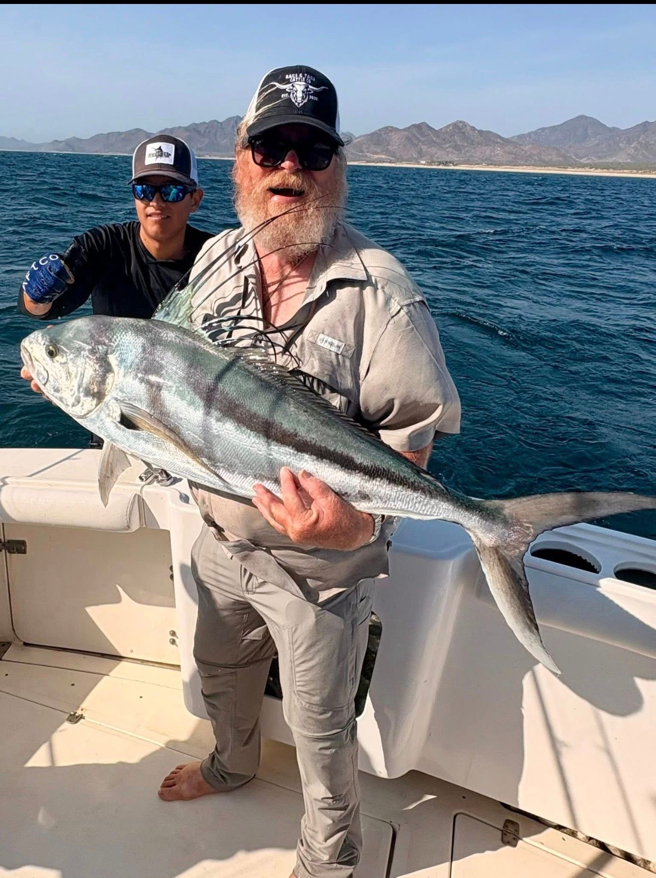 Two men on a boat holding a large silver fish with ocean and mountains in the background