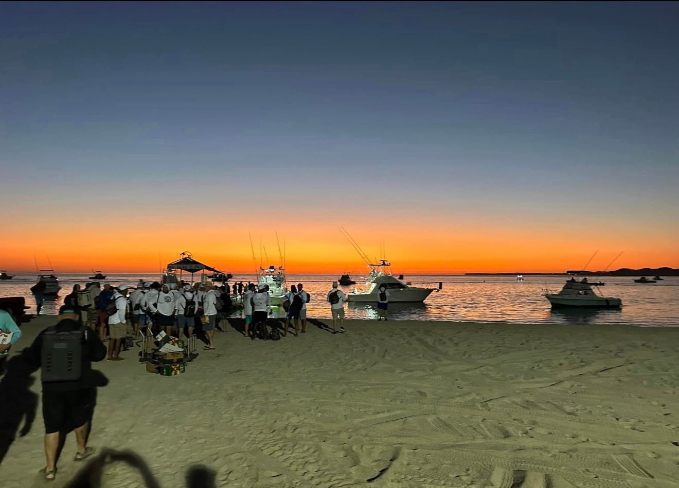 Beach at sunset with people, boats on the water, and orange sky over the horizon