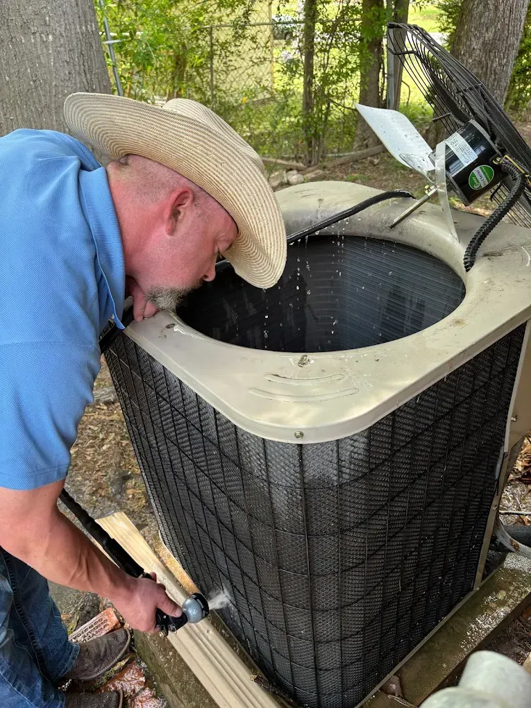 A man in a cowboy hat is cleaning an air conditioner with a hose.