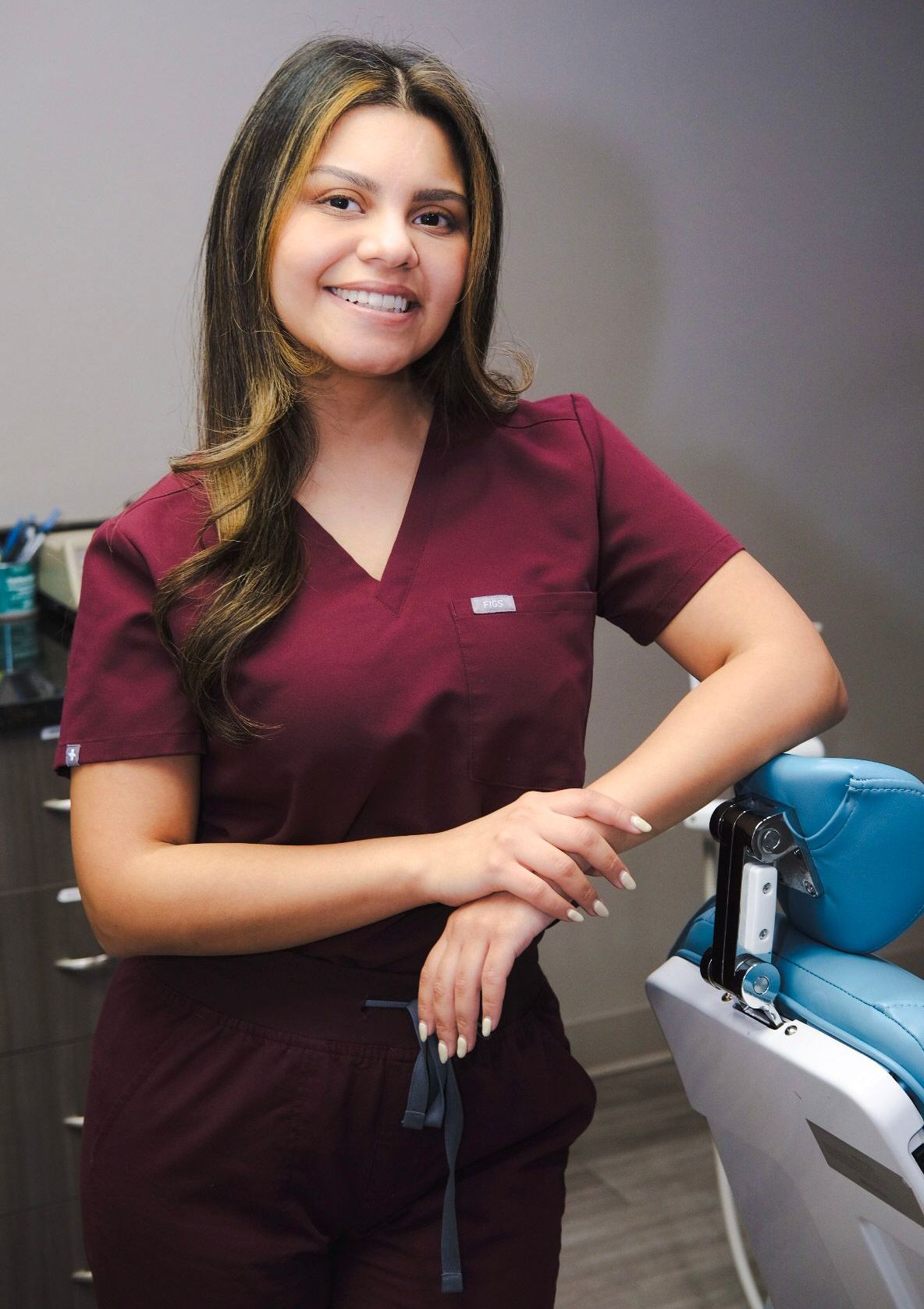 A woman in a maroon scrub suit is standing next to a dental chair.