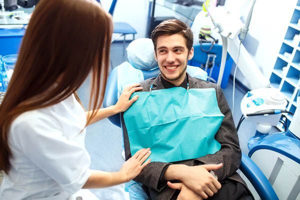 Dentist placing a bib on a patient in a dental chair; the patient smiles.