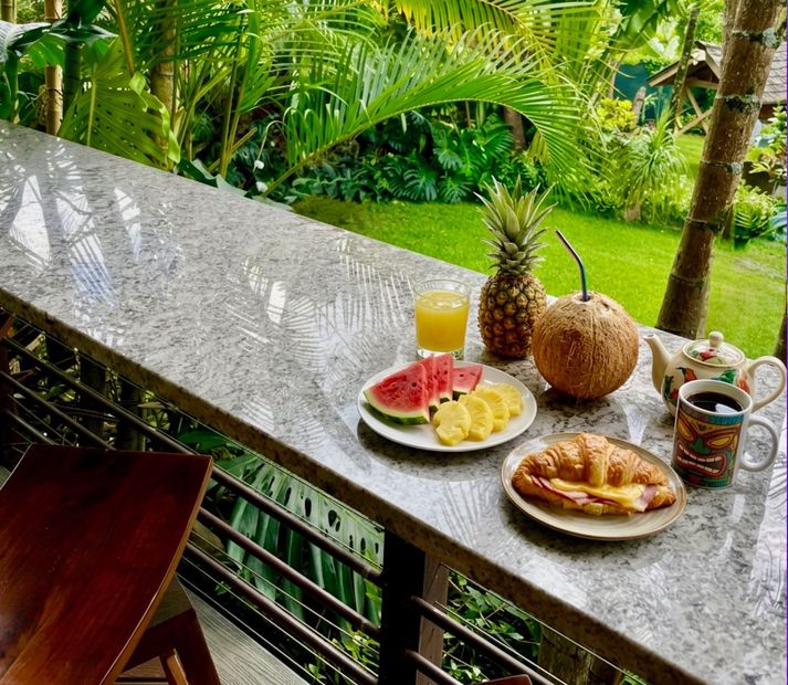 Breakfast spread on a balcony with fruit, juice, coffee, and pastries overlooking lush green garden views