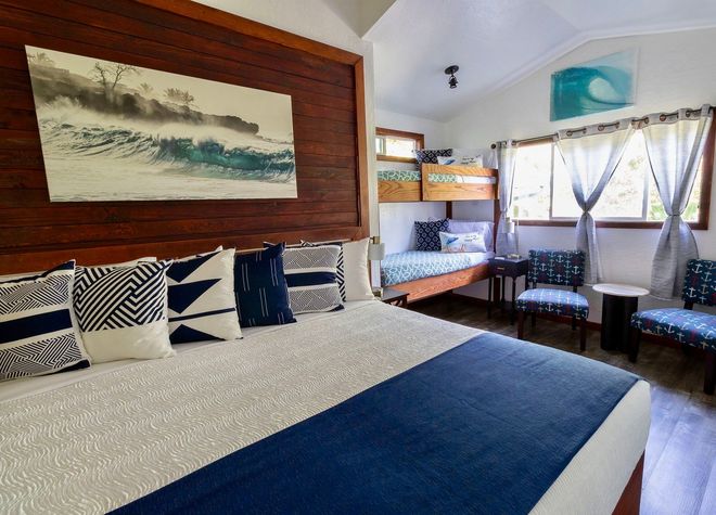 A rustic bedroom with two beds, each draped in white mosquito netting, with a lamp between them and a small window.