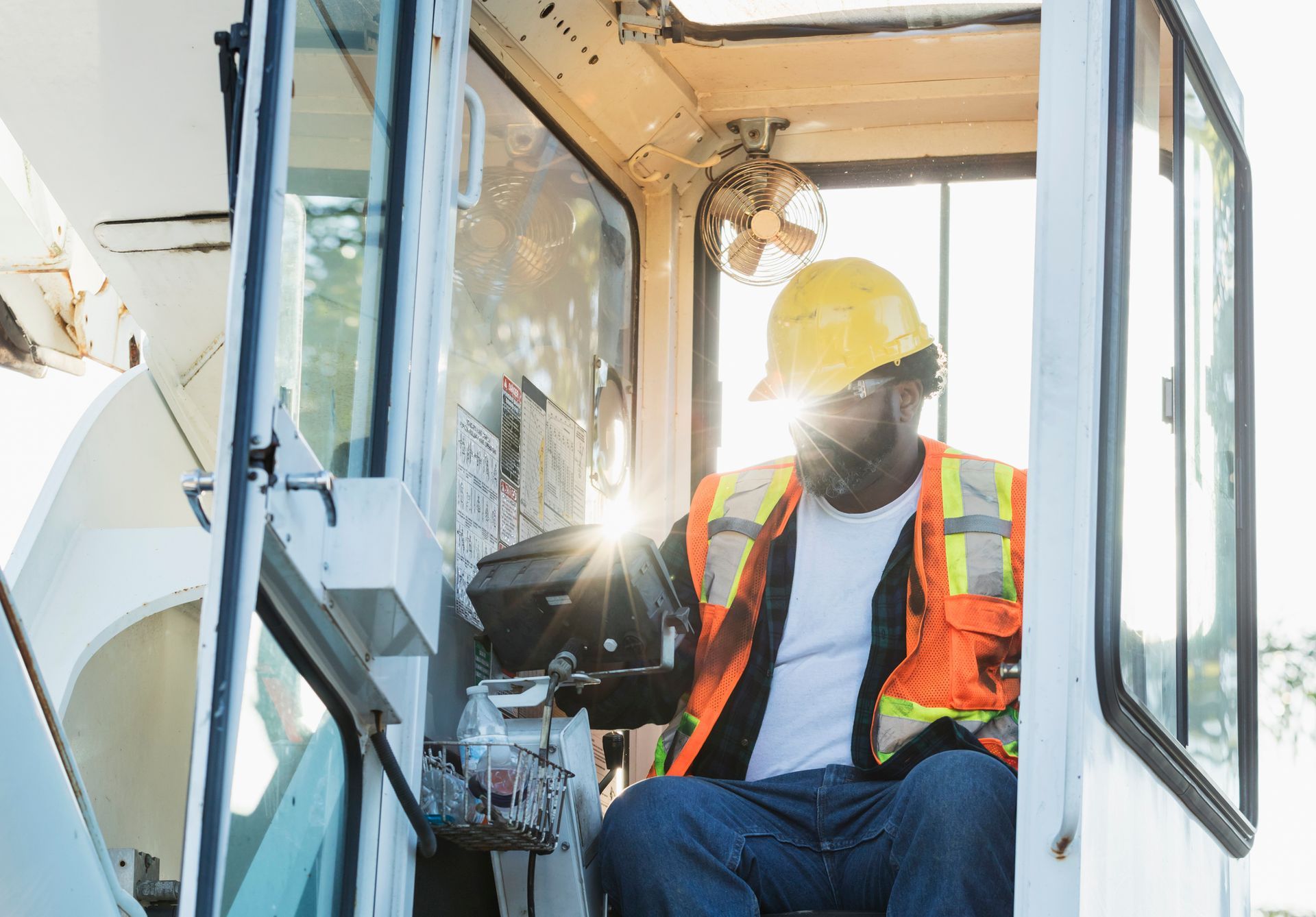 A crane operator in a hard hat and vest controls machinery inside a cabin with sunlight