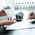 A person is holding a calculator and a pen in front of a model house.