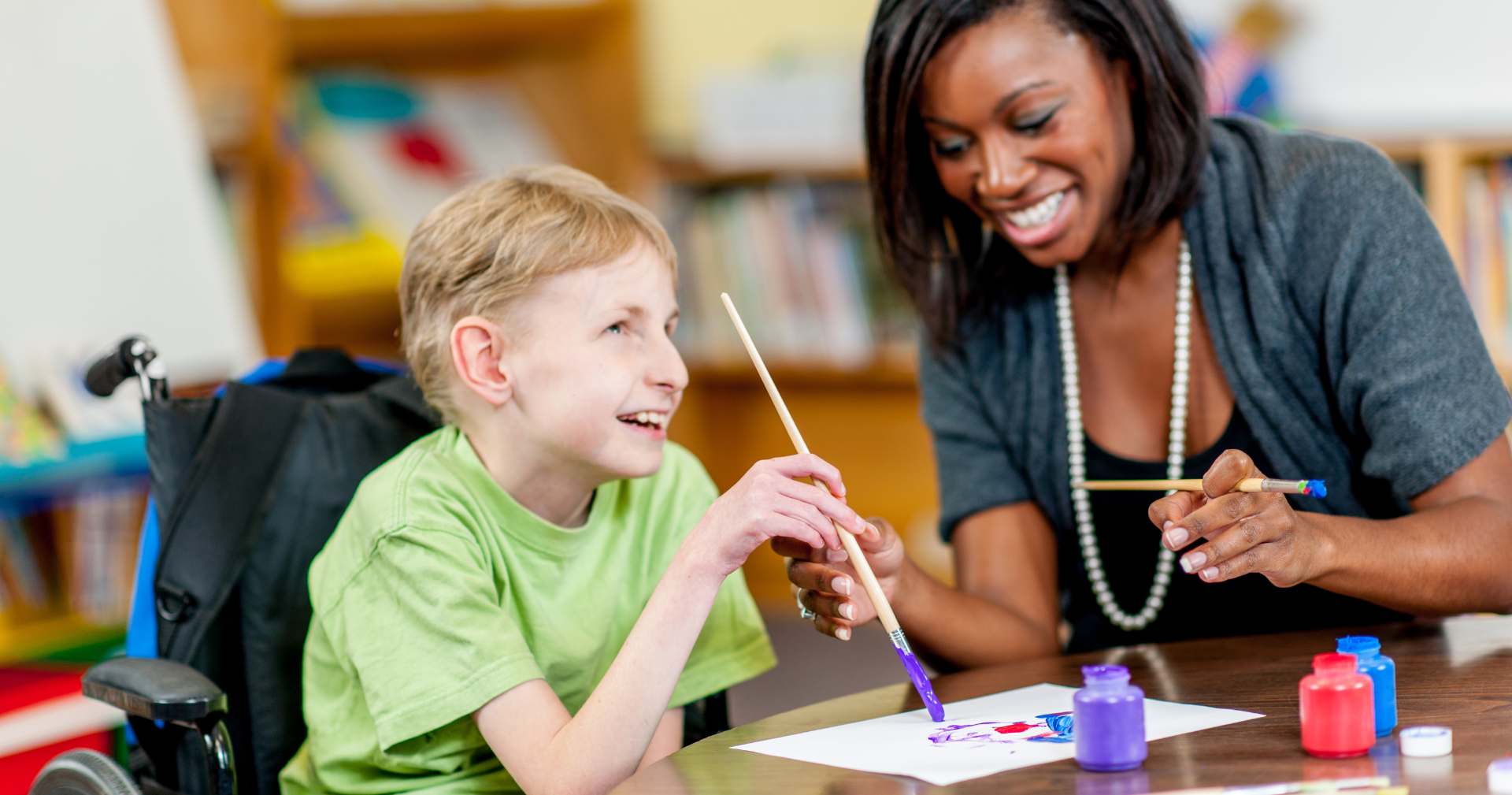 A woman is teaching a young boy in a wheelchair how to paint.