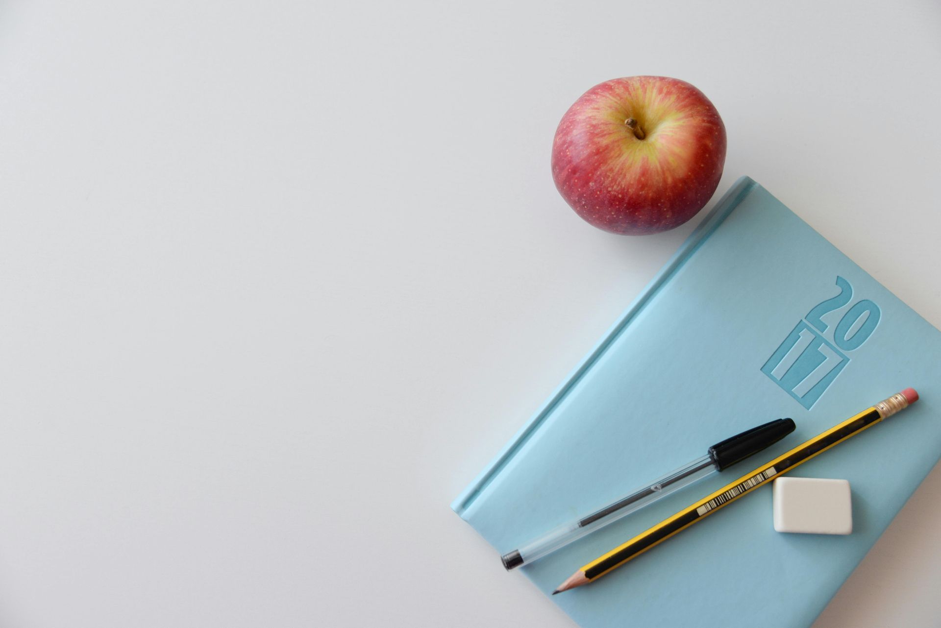 A red apple sits on top of a blue notebook next to a pencil and eraser